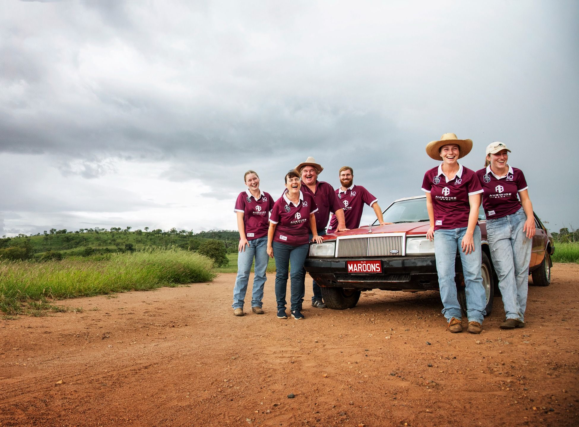 A group of smiling people in maroon shirts stand around a car on a dirt road.